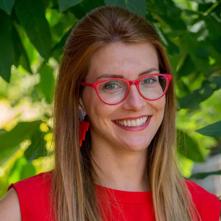 Smiling woman with red glasses and red top outdoors among green leaves.