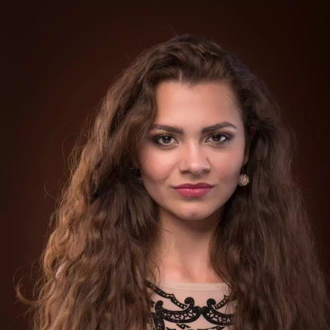 Portrait of a woman with long wavy brown hair, pearl earrings, and subtle makeup against a dark background.