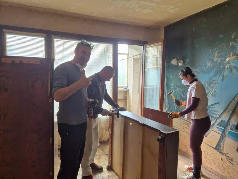 Three people renovating an indoor space; one man gives a thumbs-up while two others cut a wooden panel, all wearing gloves and masks on a sunny day interior site.